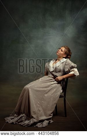 Vintage Portrait Of Young Sad Girl In Gray Dress Of Medieval Style Sitting On Chair Isolated On Dark