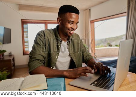 Young Adult Black Male Typing On His Laptop, Smiling. Sitting In Modern Apartment Working From Home.