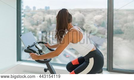 Home fitness workout woman training cardio on bike cyle with online gym class streaming. Girl biking on stationary bicycle indoor.