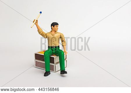 Miniature Toy Businessman Figure Sitting On A Matchbox Holding Mach Stick, On White Gray Background.