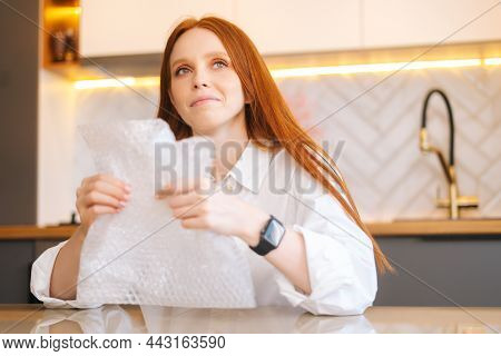 Close-up Portrait Of Smiling Attractive Young Woman Playing Pops Bubble Wrap To Calm Herself Sitting