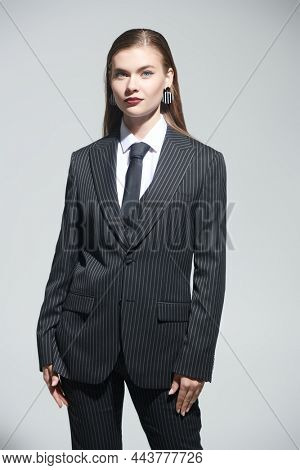 Fashion shot of a pretty young woman in a strict striped pantsuit posing smiling on a grey studio background. Impeccable look. 