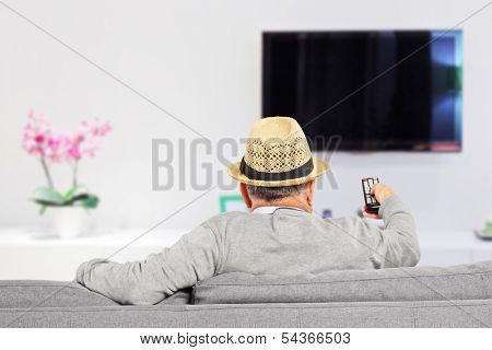 Man with hat sitting on a sofa and changing TV channels at home