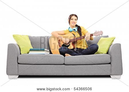 Beautiful female musician seated on a sofa playing an acoustic guitar isolated on white background