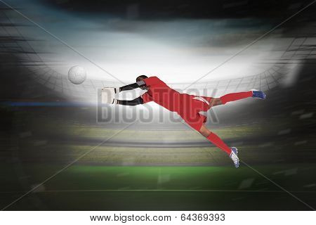 Goalkeeper in red making a save in a large football stadium with lights