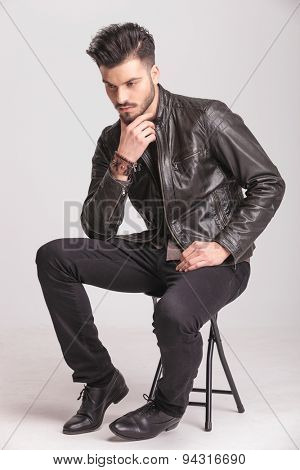Full body picture of a young casual man sitting on a chair while peting his beard, looking down.