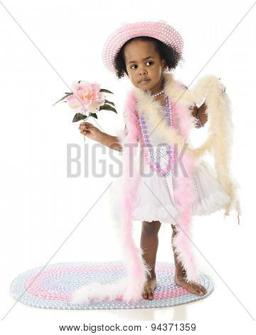 An adorable 2 year old looking in the distance as she stands in her petticoat, beads and boats.  He holds a large flower in her hand.  On a white background.