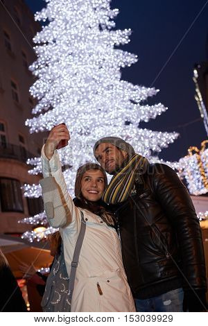 Happy young couple making selfie outdoors front of huge christmas tree.