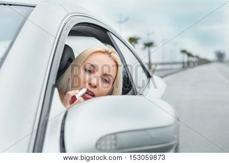 Woman apply rose lipstick looking in car mirror
