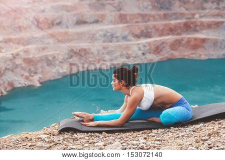 Young sporty girl wearing blue activewear stretching outdoors. Woman training yoga pose on mountain hill, calm and silent view.