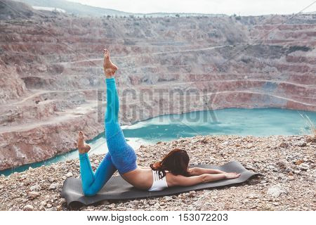 Young sporty girl wearing blue activewear stretching outdoors. Woman training yoga pose on mountain hill, calm and silent view.