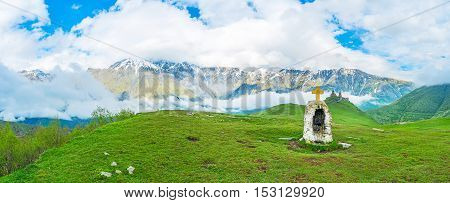 The green meadow with tiny chapel and Gergeti Trinity Church with the fluffy clouds on the background Kazbegi Georgia.
