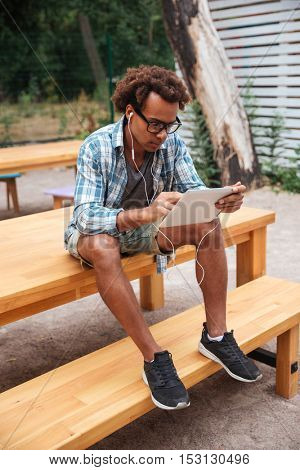 Concentrated african young man using earphones and tablet in patk