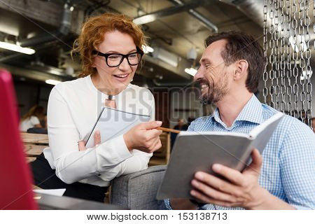 Compering results. Smiling ginger woman pointing with pencil at notes of cheerful bearded man.