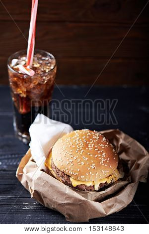 burger on a paper package served with a glass full of ice and drink. At a dark wooden background. Close view. Vertical shot