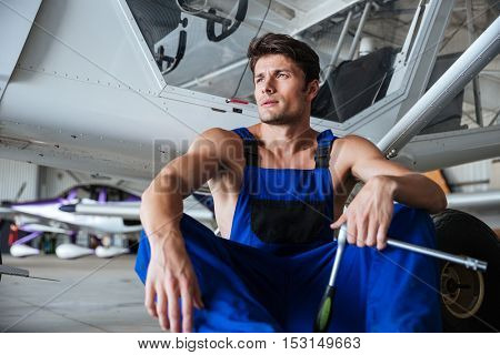 Young handsome mechanic in overall holding wrench resting at the shed