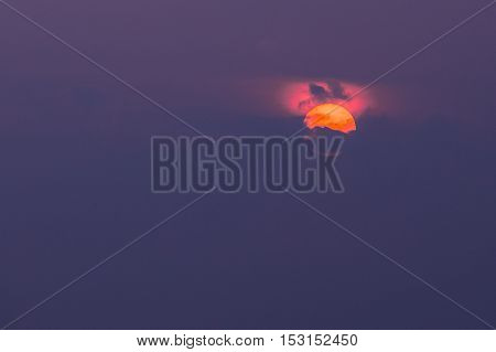 Close View Of Orange Disk Of Setting Rising Sun Partly Covered By Dark Clouds With Crimson Glow Atop, Violet Sky Background, Sunrise, Sunset, Dawn.