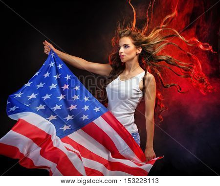 Attractive young girl holding a US flag on the flagpole. She a T-shirt and shorts with a print of the flag. Long curly hair flying. Portrait of a real patriot. Stars and Stripes. Smoke in the background.
