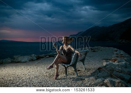 pretty sexy woman or girl with wine glass sits on bench outdoor over dark twilight sky with clouds on beach with sea or ocean water on evening natural background