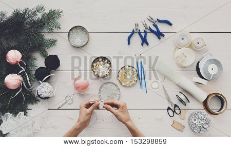 Making handmade christmas balls. Woman's leisure, tools for creating holiday decorations and garland. Top view of white wooden table with female hands holding various trinkets, decor accessories