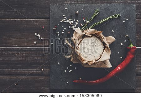 Cheese delikatessen closeup with chili pepper on black stone surface and wood. Camembert or brie circle in brown kraft paper decorated with basil, top view image