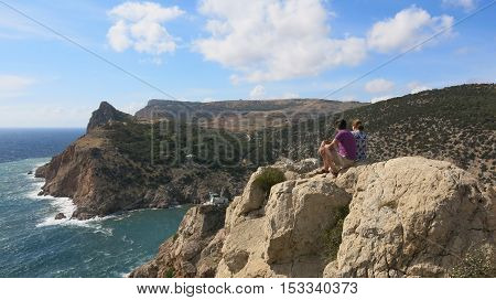 Two people sitting on the edge of a cliff and look down on the sea