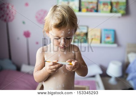 Beautiful little two year old girl at home sick with chickenpox, white antiseptic cream applied to the rash. Holding thermometer, looking at temperature.