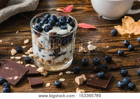 Granola with yogurt blueberries honey and chocolate bars on wooden table with ingredients.