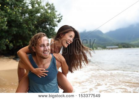 Two young people playing having fun laughing in love. Interracial couple piggybacking, boyfriend carrying Asian girlfriend doing piggyback on hawaiian beach in Kauai, Hawaii travel vacation.
