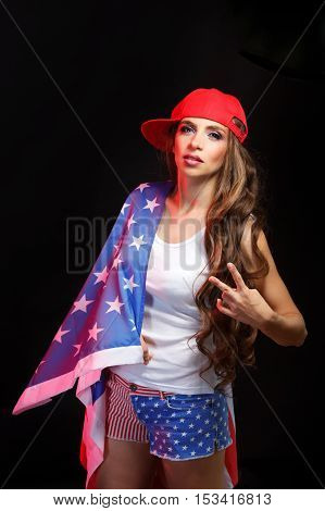 Young attractive girl in a baseball cap and sunglasses holding a US flag. She a T-shirt and shorts with a print of the flag. Long curly hair. Patriot country. She shows the victory.