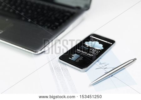 business, technology forecast and meteo concept - close up of smartphone with weather cast on screen, laptop computer and pen on office table