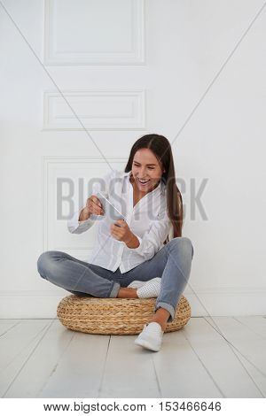 An attractive young Caucasian woman in casual clothing is radiant with the joyful emotions while playing on tablet and sitting on a hassock