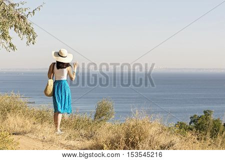 Young woman (brunette) in a blue skirt and hat looks at sea. Selective focus.