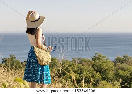 Young woman (brunette) in a blue skirt and hat looks at sea. Selective focus.