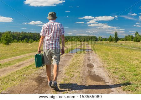 Mature man wearing blinders shirt and hat walking on a summer country road with green suitcase