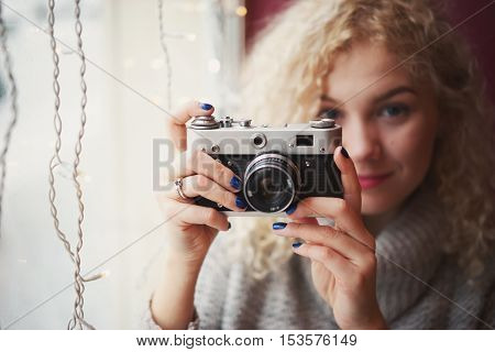 Young blond curly female in warm sweater with old film camera smiling and shooting a photo in the cafe winter city outside the window