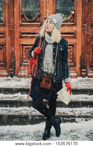Young blond curly female in warm clothes with old film camera and map standing. Wooden doors with handles on the background winter
