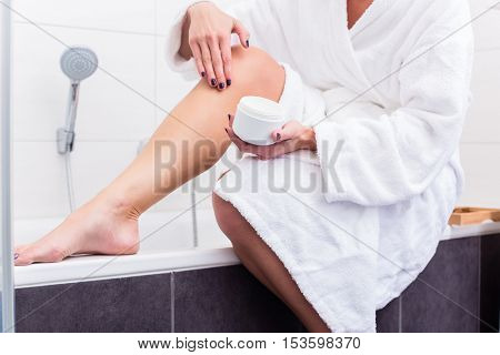 Woman in bathroom sitting at edge of bathtub putting on lotion on legs