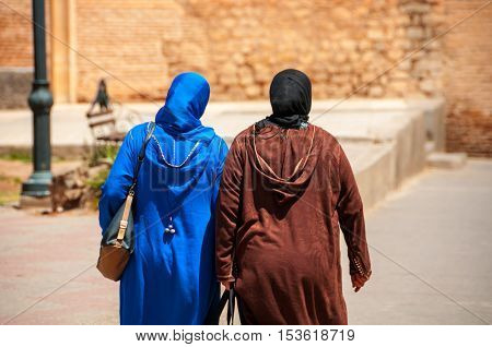 Two moroccan women seen from the back dressed in typical blue and brown djellaba walk down the streets of Marrakesh Medina, Morocco