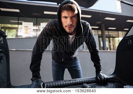 Criminal young man in black hoodie and gloves standing and looking at car trunk