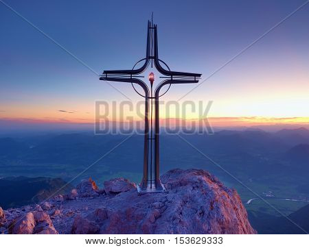 Steel Crucifix At Mountain Peak In Alps. Sharp Rocky Summit, Daybreak Sun In Sky.