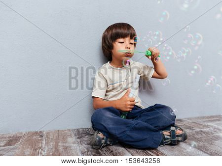 Full length of cute little boy sitting and blowing soap bubbles over gray background