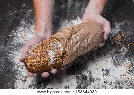 Warm fresh rye whole grain bread, wrapped in craft paper. Healthy baking and cooking concept background. Hands carefully hold loaf on wooden table background, sprinkled with flour. Soft toning