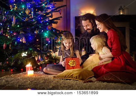 Young happy family of four unwrapping Christmas gifts by a fireplace in a cozy dark living room on Christmas eve