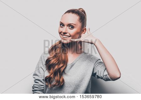 Call me! Attractive young woman gesturing telephone near face and smiling while standing against grey background