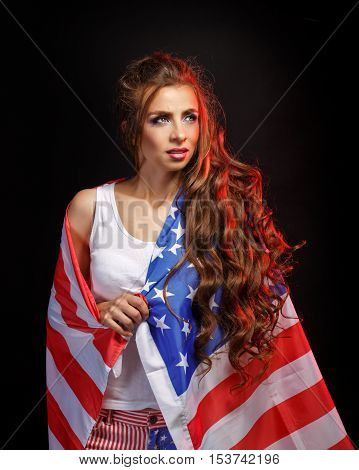 Young attractive girl wrapped in a US flag. She a T-shirt and shorts with a print of the flag. Long curly hair. Portrait of a real patriot. Stars and Stripes.