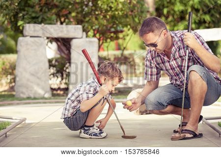 Sharing with golf experience. Cheerful young man teaching his son to play mini golf at the day time. Concept of friendly family.