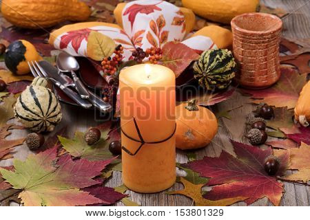 Selective focus of a close up view of holiday candle burning with autumn dinner setting with real gourd decorations leaves and acorns on top of rustic wood