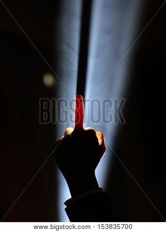 dark silhouette of human male hand with raised middle finger in spotlight or backlight light with gesture on black background with dramatic projector shine ray or beam