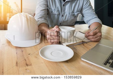 engineering useing laptop computer note someting with pen at notbook and holding a cup of coffee on wooden table